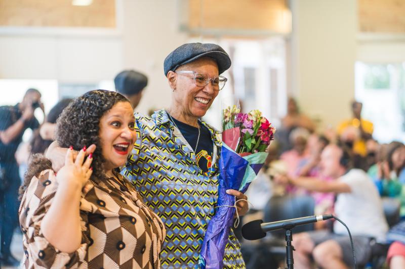 Black woman with long curly hair (left) smiling next to a Black woman with a hat on who is holding a bouquet of flowers (right). Black woman with long curly hair (left) smiling next to a Black woman with a hat on who is holding a bouquet of flowers (right).