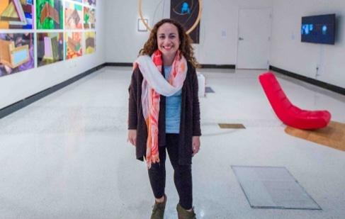 : Amanda Cachia is a white, short-statured young woman with shoulder-length, brown, curly hair. She is wearing a blue shirt, a purple cardigan, black leggings, and a white and pink colored scarf draped over her neck and chest. She is standing inside an art gallery with white walls, to her left is a collection of colorful, rectangular paintings, and to her right is a red sofa chair and tv on the left wall.