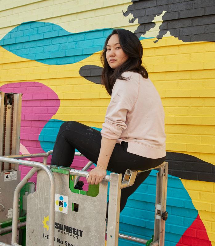 Asian women on scaffolding in front of mural she is painting. 