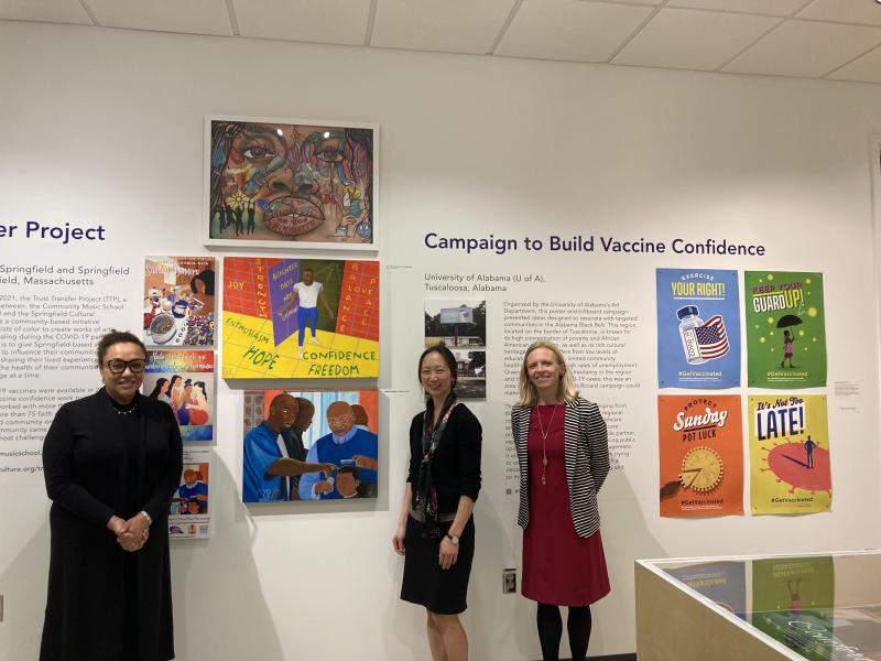 Woman in black dress with glasses and hair pulled back next to woman in short black dress with long black hair and woman with blond hair in purple dress and striped blazer standing in front of artwork in a museum.