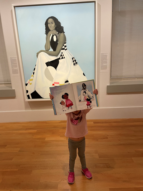 Little girl with grey pants, a pink shirt, and pink shoes holding a picture book up in front of the Michelle Obama painting. 
