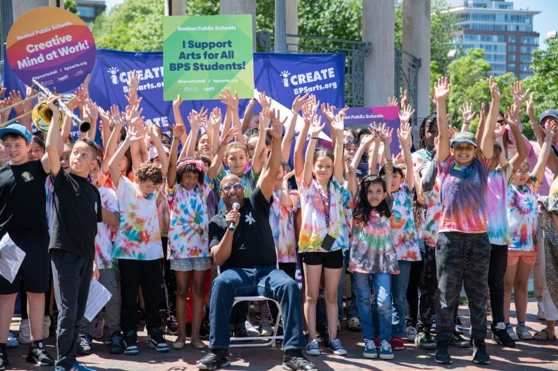  A group of children celebrate the arts in Boston Public Schools. They are outside with their hands raised high, some holding signs that say "iCreate" and "Creative Mind at Work." Many are wearing tie-dyed t-shirts and some hold musical instruments. 