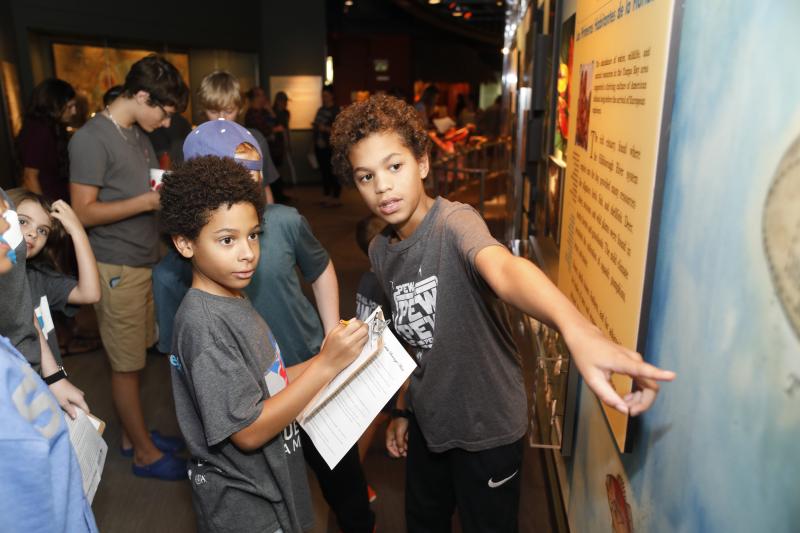 Two boys engage in an art exhibit at a museum. In the foreground, one child pointing at an information board while another take notes. 