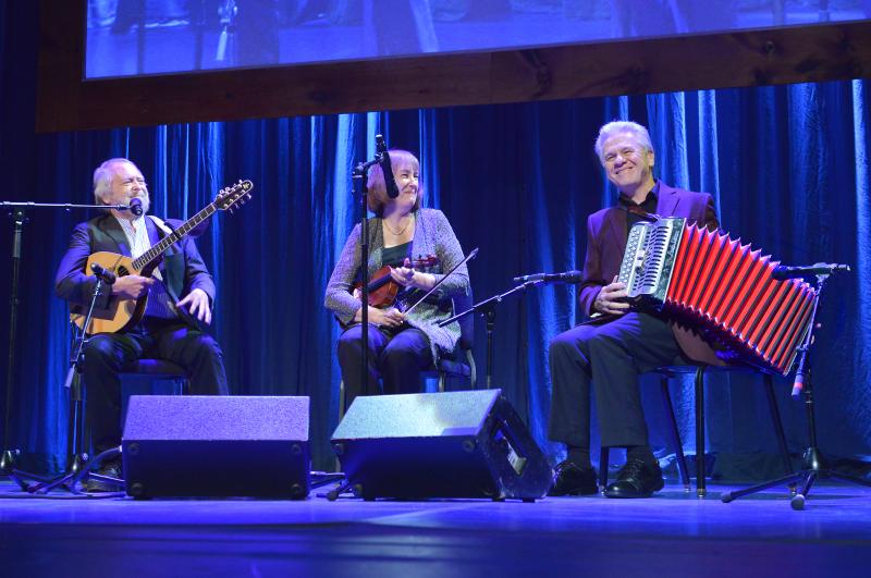 Heritage Fellows Mick Moloney Liz Carroll and Billy McComiskey Photo by Tom Pich Three people play on stage with a blue background. A man with a banjo and woman with a fiddle look at a man playing the button accordian