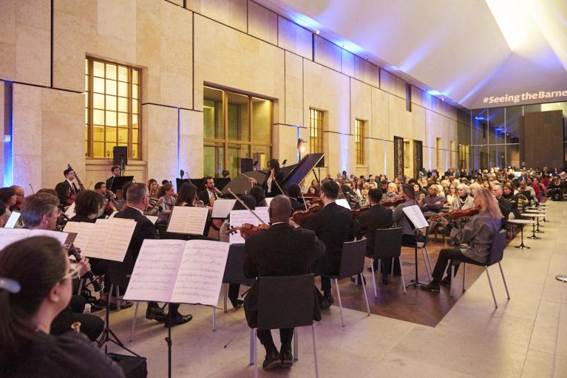 a Black woman conducts surrounded by members of the orchestra playing their instruments