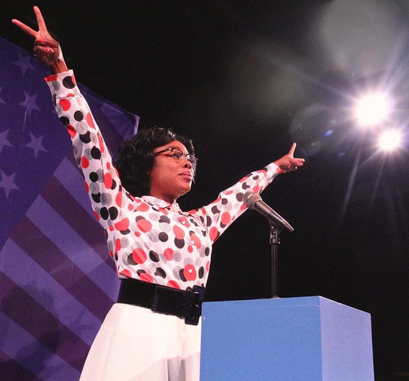 Black woman with glasses, wearing a polka dot blouse, black belt, and white skirt as she holds up with peace sign on both hands.