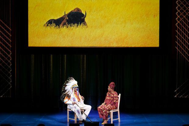 A man in Native American regalia sits on a stage across from a woman. An image of a buffalo is on the screen behind them.