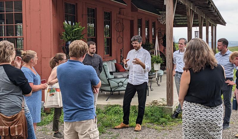 People stand in a circle outside listening to a man in a white shirt and black pants. There is a red barn in the background.