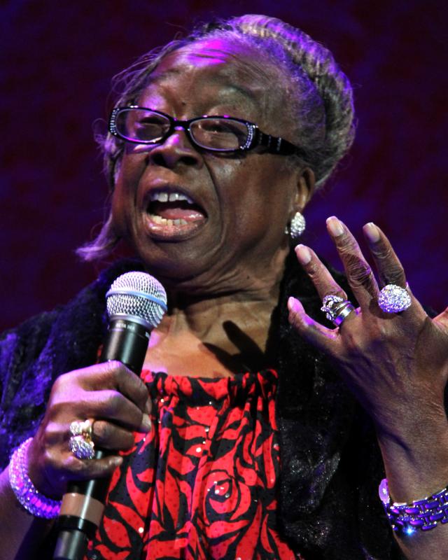 A woman with glasses and sparkly jewelry sings into a microphone