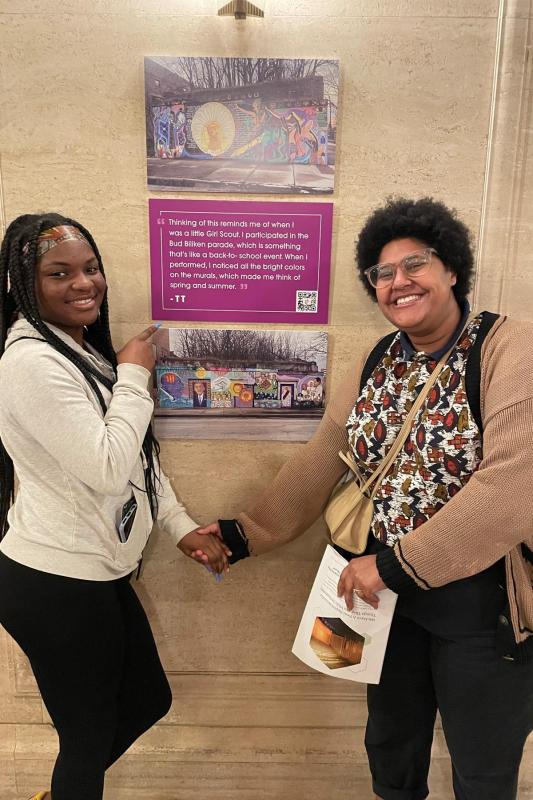 Black teen female (left) with braids in her hair and wearing a grey hoodie and black pants, holds hands with a Black woman (right) wearing glasses, a neutral print top, tan cardigan, and black pants. The Black teen female (left) is pointing her fingers at a brown wall exhibition that has a poem written with purple and white colors.