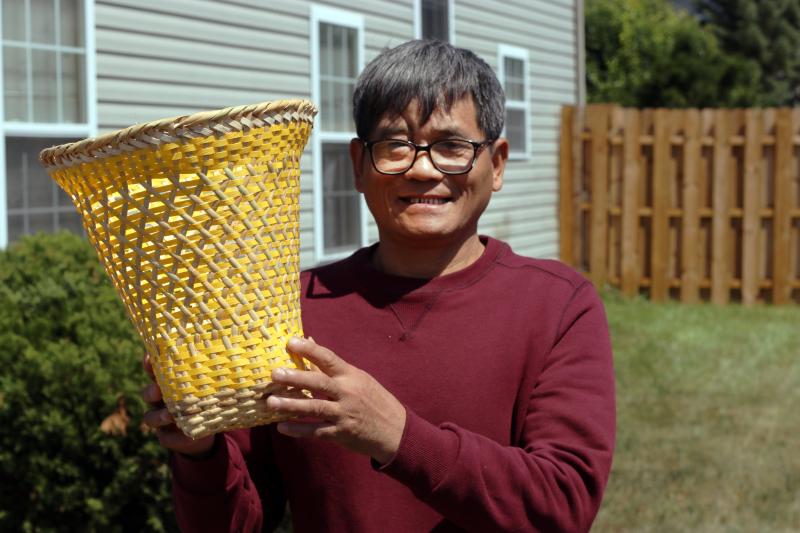 A smiling Burmese Chin man wearing glasses and a red sweater holds up a handmade basket. 