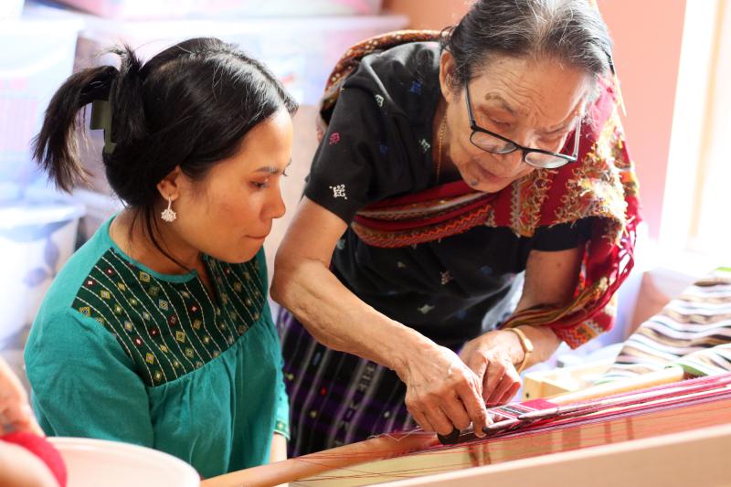 An older Asian woman stands over a loom with a younger Asian woman, teaching her to weave