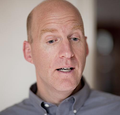Chris Downey is a white man in his fifties with no hair on top of his but hair on the sides his hair color is a dark blond. He is wearing a gray button-down shirt with white buttons. Downey appears to be talking mid-sentence in this photo and is gazing downward. Downey is in the center foreground and the background is blurred.