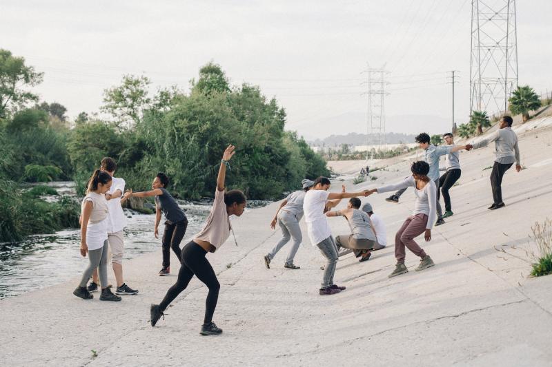A group of dancers in neutral colored clothing perform on concrete next to a river with lush vegetation. 