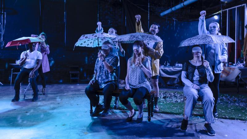 Four individuals sit in chairs holding umbrellas and wearing blindfolds. Behind them, four people stand with their hands raised, prepared to drop something onto the umbrella. 