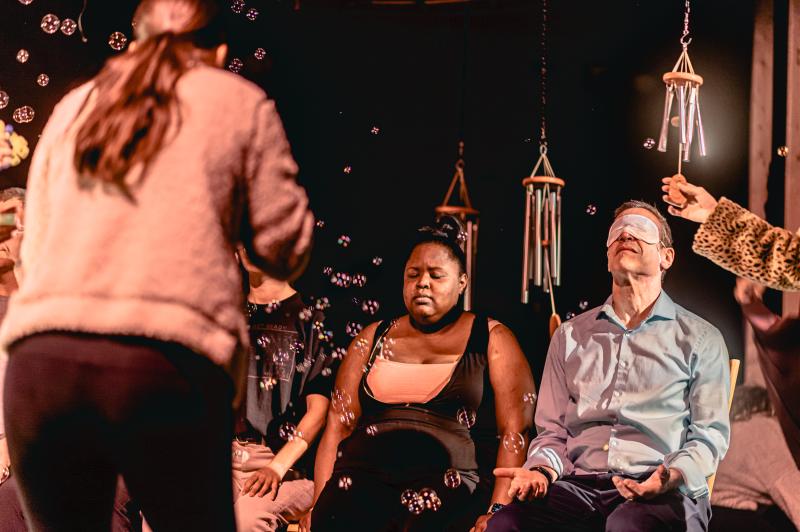 Two people sit on a stage below windchimes as a third person blows bubbles towards them