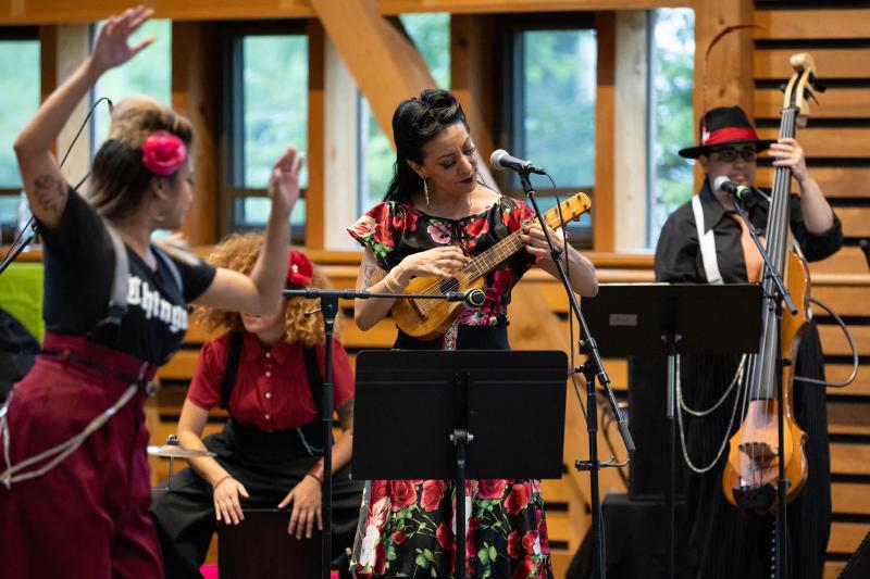 A woman wearing flowery dress and a man with a wide-brim hat playing string instruments on stage with woman dancing in front of them out of focus.