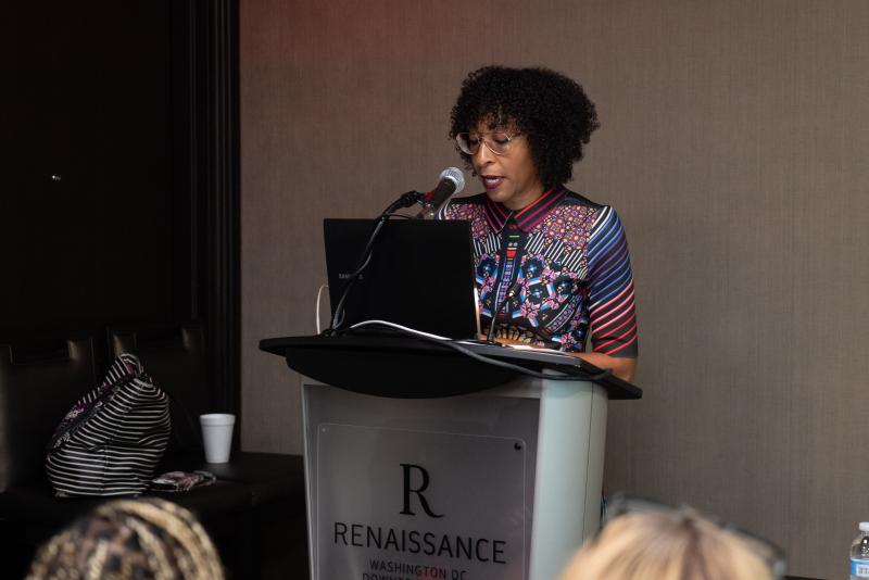 Black woman wearing glasses and a colorful shirt with medium-length black hair speaking at a podium on stage.