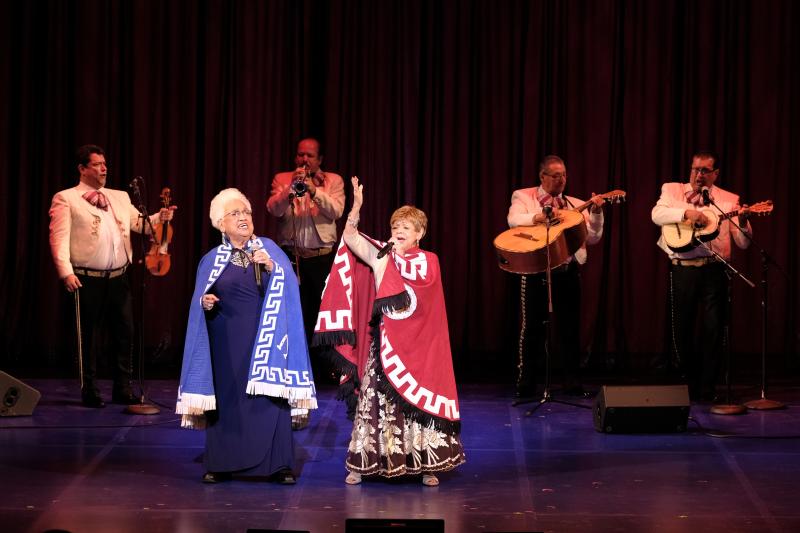 Two older women-one wearing a blue cape and one wearing a red cape-sing on stage with a mariachi band in the background