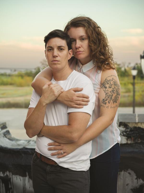 Two white people, one with short cropped brown hair and the other with long curly brown hair, look directly at the camera, their intertwined pose both protective and relaxed.