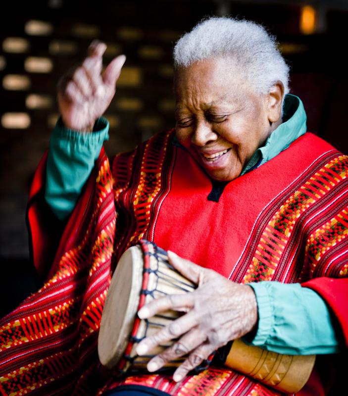 An older woman sits playing a talking drum. She is wearing brightly colored clothing and smiling as she gets ready to strike the drum again in song.