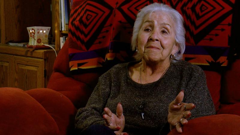 Woman with gray hair wearing dark sweater sitting on red, pillowed chair with Native-designed blanket behind her head.
