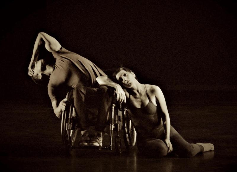 Black and white photo: dancer in a wheelchair wearing a tshirt and pants and dancer on right is in a leotard and leaning on the left dancer's wheelchair.