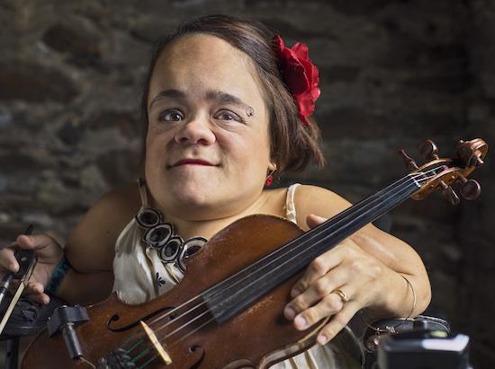 Gaelynn Lea is a white woman with medium-length brown hair and brown eyes. She has a red flower pinned to the right side of her hair, her left eyebrow is pierced, and she is wearing a white dress with dark round beads at the top, while sporting a gold ring and bracelet on her right hand and arm. Gaelynn is holding a brown violin in her left hand and a violin bow in her right hand. She is seated in an electric wheelchair in front of a dark colored backdrop.