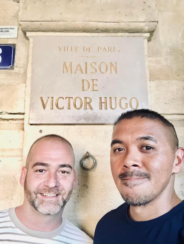 Man (left) wearing a tan and white striped shirt and man (right) wearing a dark blue shirt standing in front of a tan building that has the verbiage "VILLE DE PARIS MAISON DE VICTOR HUGO"