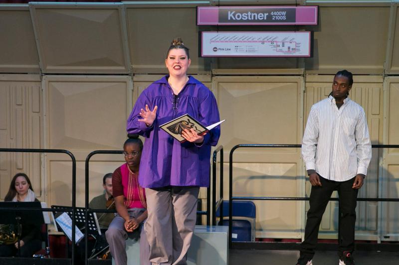  On stage: White woman (center stage) wearing a blue jacket and holding a book and on the left, a Black man with a buttoned down white and black shirt staring at the woman center stage. On the left is a Black female wearing glasses and a purple top, sitting on a grey box. Kostner subway prop (black, white, and purple) is hanging above the stage.