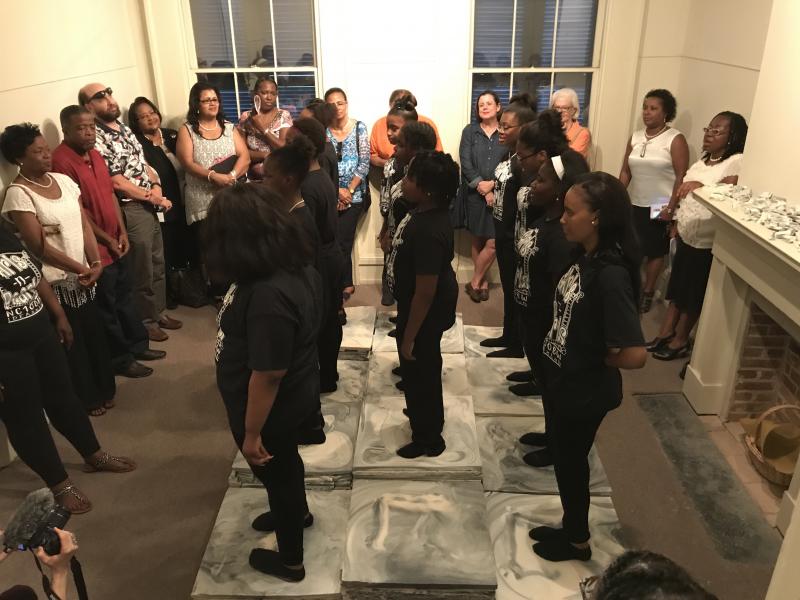 Young women standing in center of small room as part of a performance surrounded by adults watching.