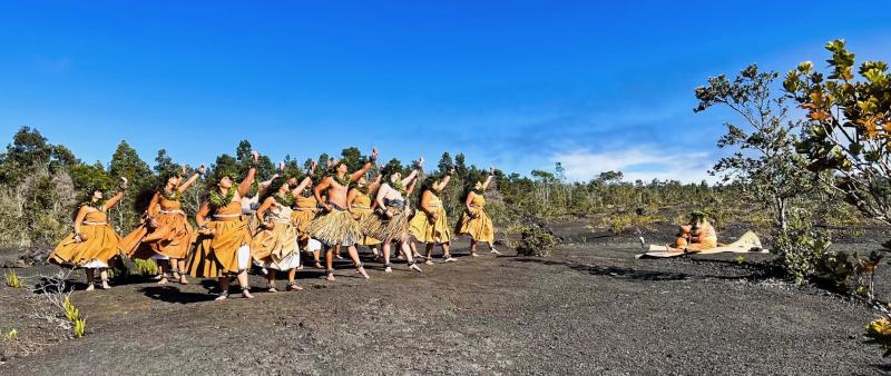 A group of hulu dancers perform outside with a musician sitting in front of them.