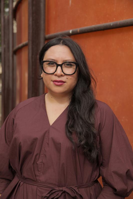 Portrait of woman with long dark hair wearing black-rimmed glasses and wearing a purple shirt. 