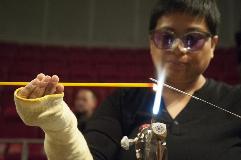 Person wearing goggles holds a yellow glass rod over open flame