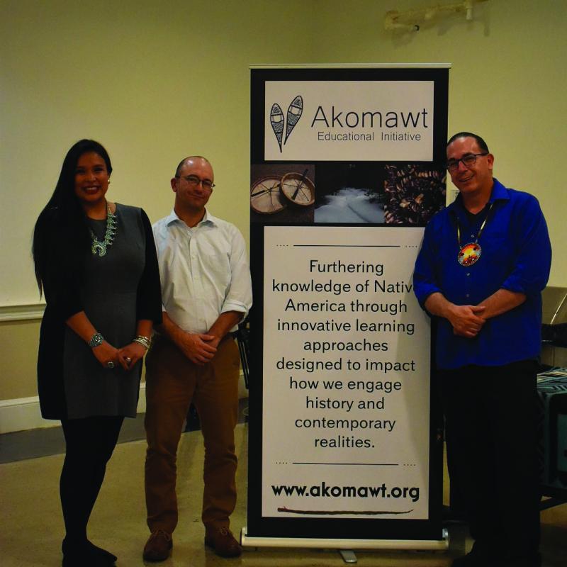 A woman in a blue dress, a bald man with a white shirt and glasses, and a man in a blue shirt with glasses standing in front of a sign.