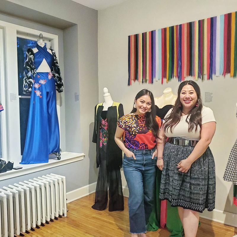 Two women pose together. Behind them are stands with clothing displayed and a colorful banner on the wall