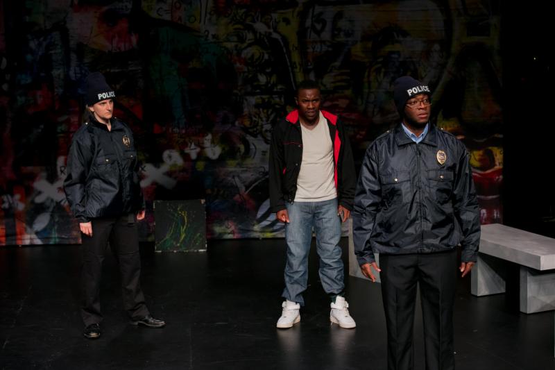 On stage: White actress (left) in a black police outfit glaring at a Black actor (center) wearing jeans and a black and pink hoodie, who is glaring at a Black actor (right) who is wearing glasses and a black police outfit. On stage: White actress (left) in a black police outfit glaring at a Black actor (center) wearing jeans and a black and pink hoodie, who is glaring at a Black actor (right) who is wearing glasses and a black police outfit.