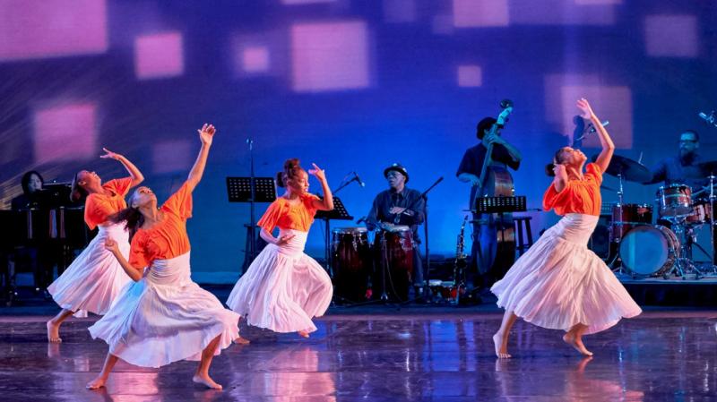four dancers in orange tops and white skirts perform. There is also a band playing upstage.