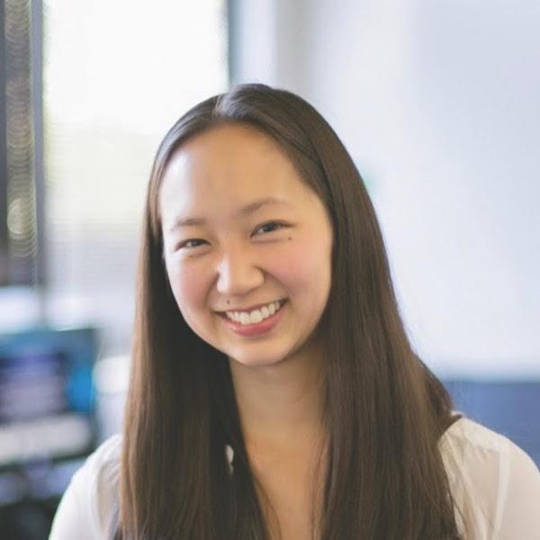 a headshot of Jenn Chang, who is an Asian-American woman with long dark hair parted in the center and falling over shoulders. She is smiling. The background behind her is blurred.