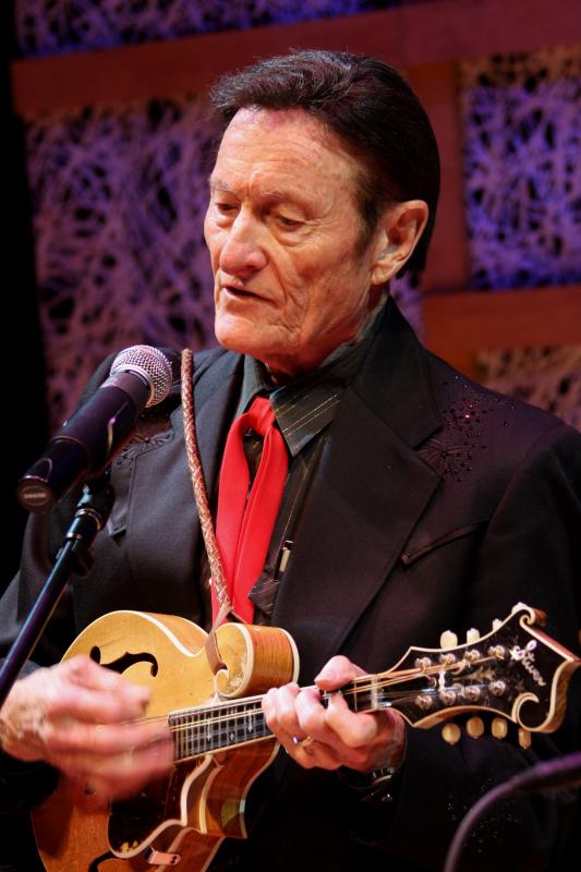 An older man with dark hair, black shirt and coat, and a red tie sings into a microphone and plays the mandolin 
