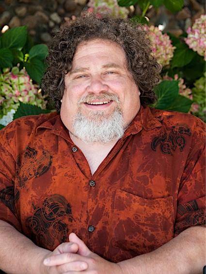 James LeBrecht is a white man in his sixties with curly brown hair and a gray and white goatee the sides of his face are clean shaven. He is smiling, looking directly at us and is holding his hands together. James is wearing a short-sleeved, red, button-down shirt with black accents. Large flowers and green leaves are displayed within the background.