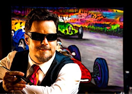 John Bramblitt is a white man in his forties. He has short, dark brown hair; he is wearing black rectangular sunglasses and has an anchor-style beard and mustache. John is wearing a white button-up, collared shirt with a dark blue vest paired with a bright red-orange tie. He is holding a paintbrush in his left hand while being seated. In the background is a single large painting out of focus placed in front a black wall.