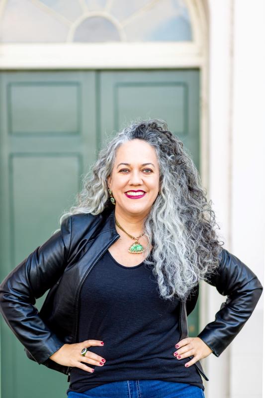 A woman with long grey curly hair stands with her hands on her hips. She wears a black shirt and black leather jacket