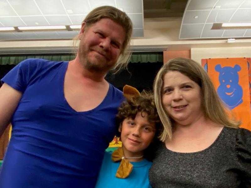 A family photo of a man with his hair in a ponytail and blue shirt; a male child with curly hair and a blue shirt; and a woman with shoulder length hair and a grey shirt. A family photo of a man with his hair in a ponytail and blue shirt; a male child with curly hair and a blue shirt; and a woman with shoulder length hair and a grey shirt.