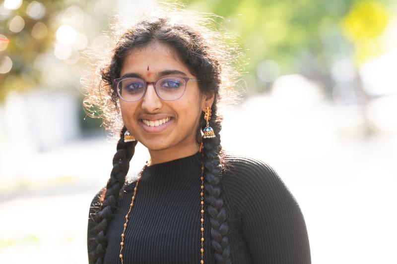 A young South Asian woman smiles, wearing a black turtleneck, long necklace, and dangling earrings. She has glasses and two long braids.