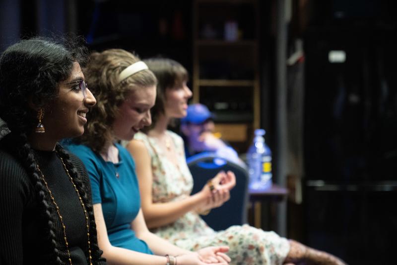 A South Asian woman wearing her long dark hair in two braids, who wears glasses and a black turtleneck, sits backstage with two other young woman visible next to her