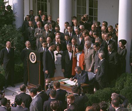 Large group of men and women surrounding President Johnson sitting at a small table signing the legislation