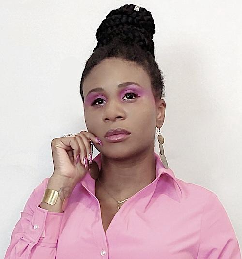 Lachi, a beautiful blind black woman, poses with a thoughtful look. She is wearing a pink shirt, gold jewelry, and pink eyeshadow, and her hair is done up in braids.