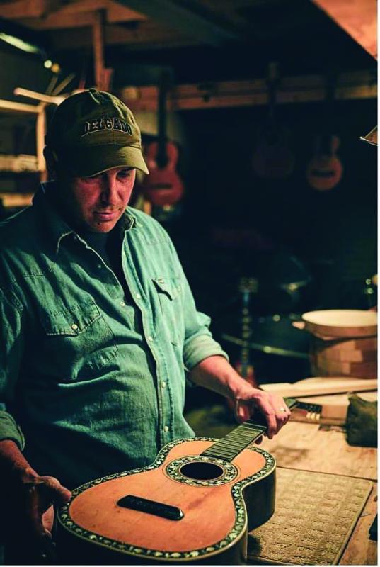 Man wearing a baseball cap and blue denim shirt looking down at a guitar. 