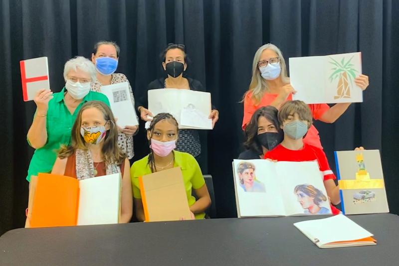 A group of people of eight people of many ages in masks hold up colorful, creative handmade books to the camera.